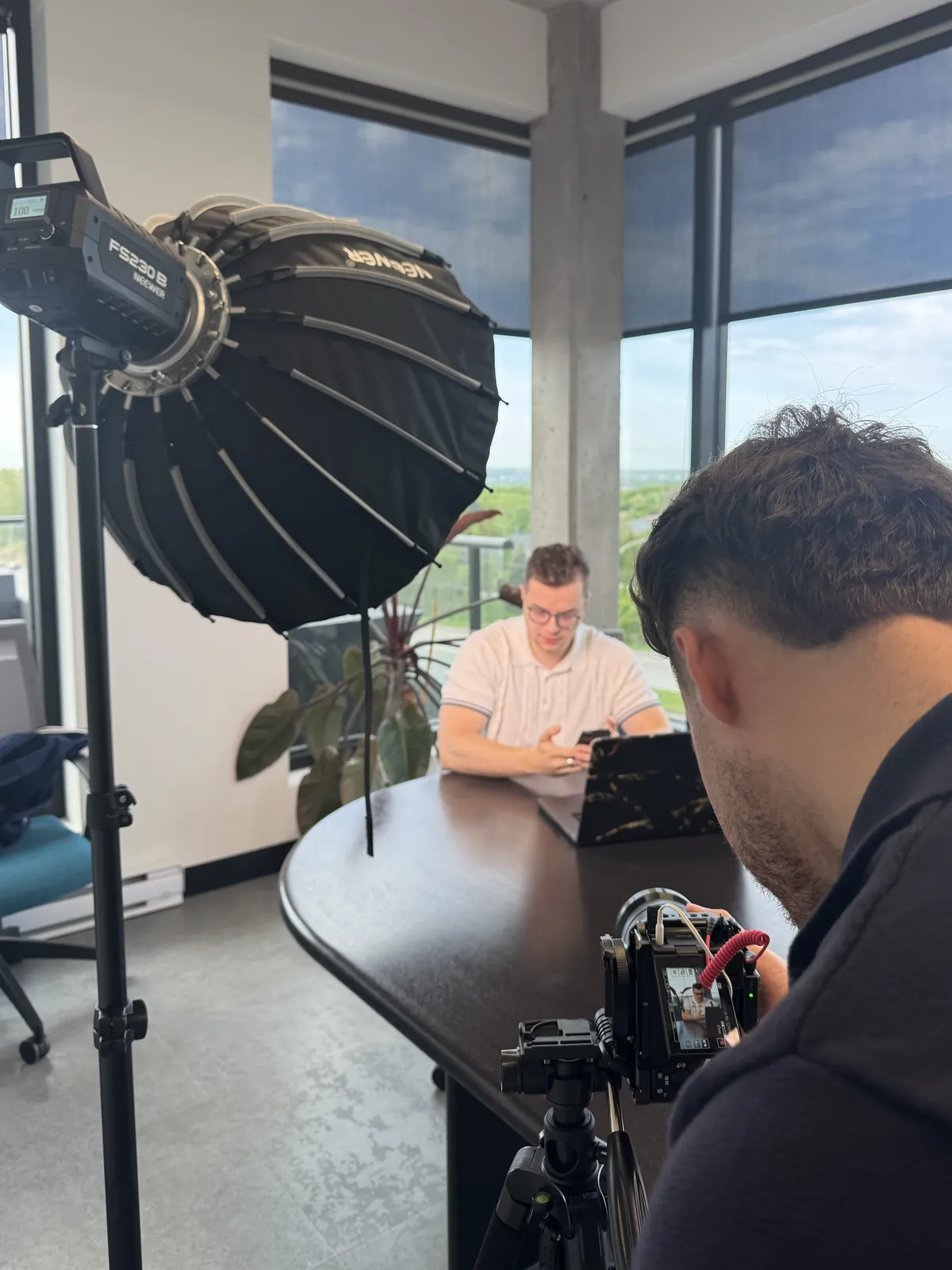 Séance de tournage Nadeauprod : un softbox monté sur pied éclaire un client assis à une table avec son ordinateur, pendant qu'un caméraman le filme au premier plan avec une caméra sur trépied.